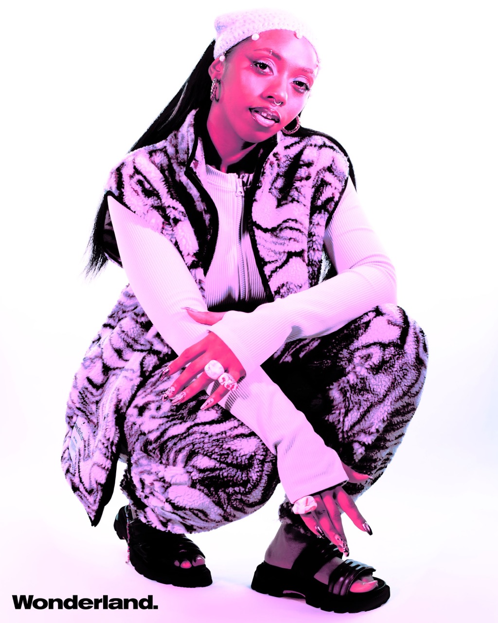 Young adult woman poses in a white photo studio wearing a patterned vest, long-sleeve top, knit hat, rings, platform sandals and Wonderland branding.