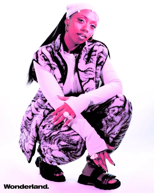 Young adult woman poses in a white photo studio wearing a patterned vest, long-sleeve top, knit hat, rings, platform sandals and Wonderland branding.