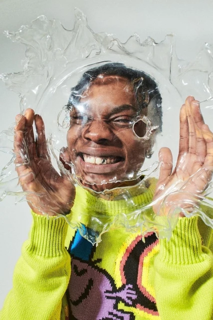 Close-up portrait of a Black adult male smiling in a neon-green sweater, wearing a clear plastic dome-shaped hat in a studio setting.