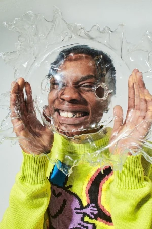 Close-up portrait of a Black adult male smiling in a neon-green sweater, wearing a clear plastic dome-shaped hat in a studio setting.