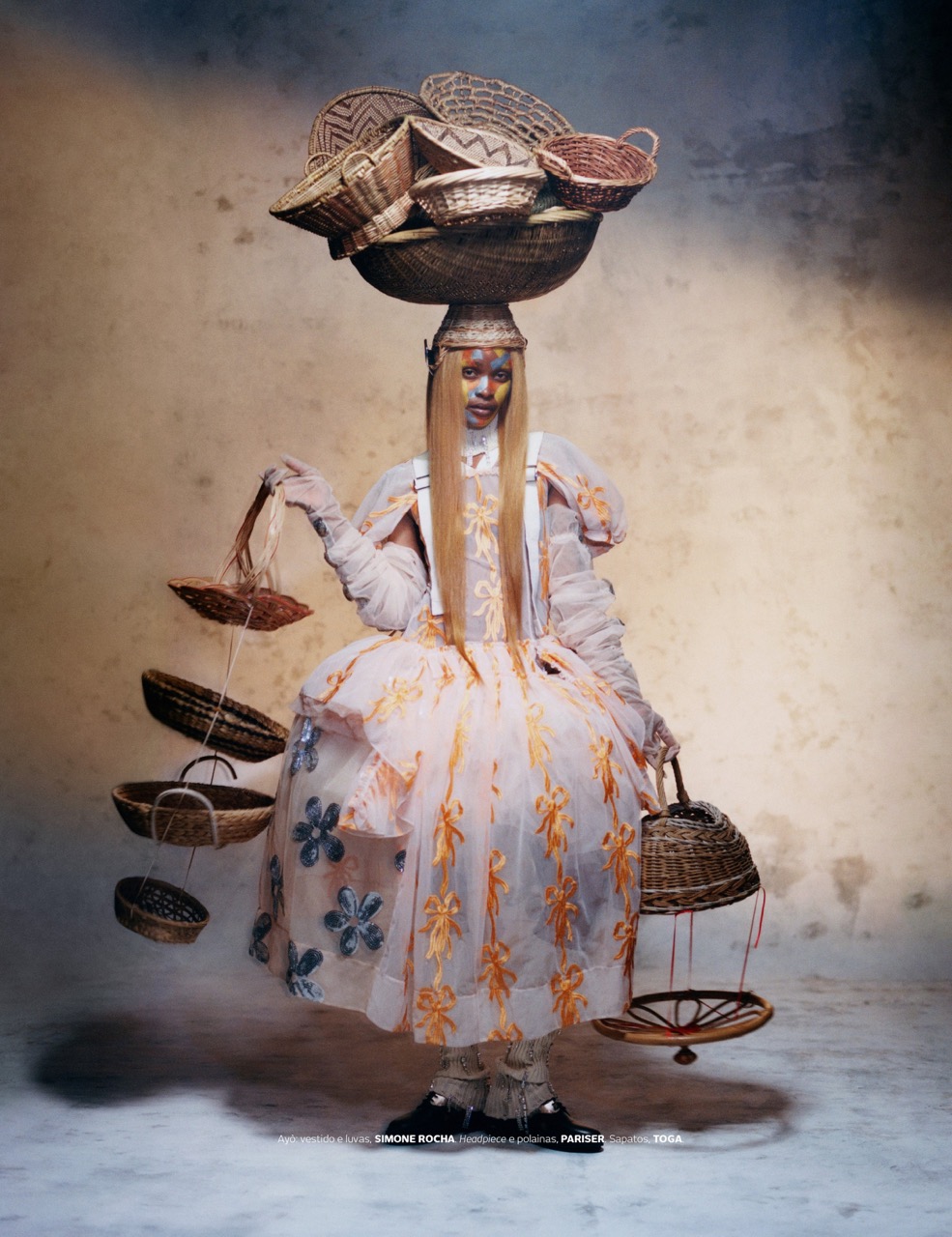 Fashion shot of a person in a flower-patterned dress balancing a large basket of smaller baskets on their head in a studio setting.