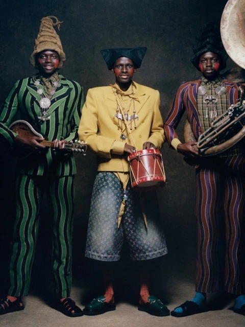 Three dark-skinned men in striped suits and hats pose with musical instruments, including a drum, mandolin, and tuba against a dark backdrop.