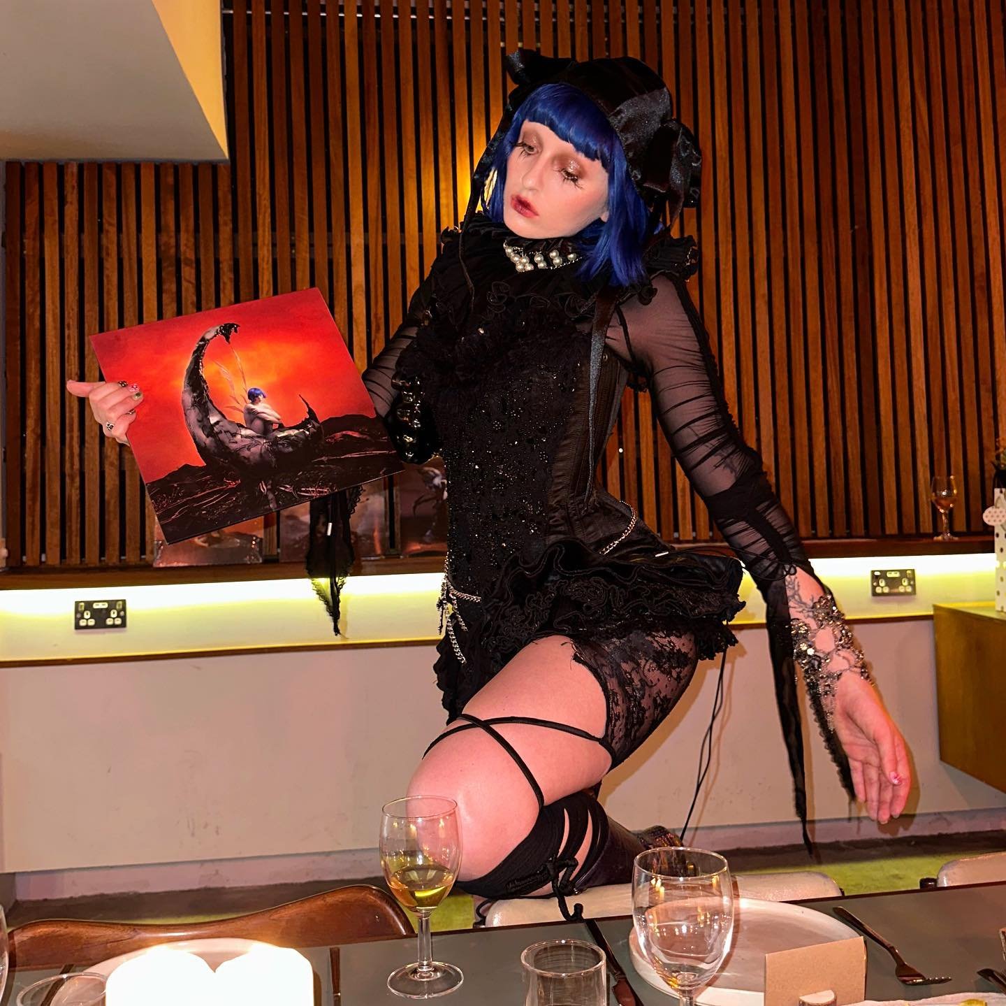 Adult woman in a black outfit with blue hair kneels at a table, holding an art print with orange and black tones in an indoor environment.