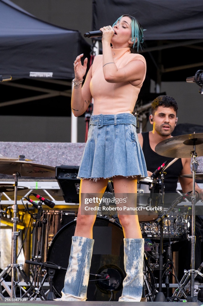 Singer performing live on stage with a microphone, wearing a light pink top, denim skirt and boots with a drummer in the background.