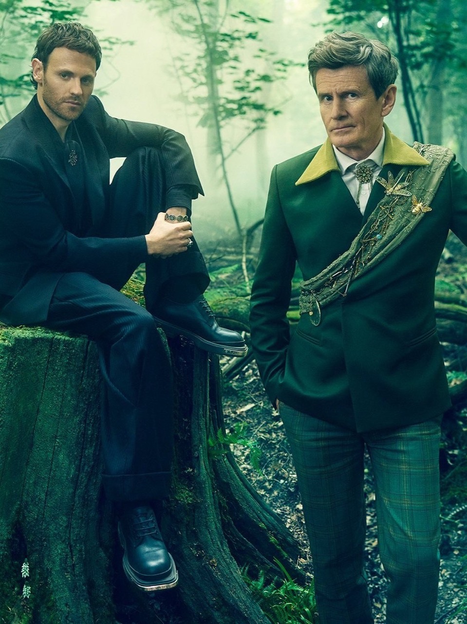 Two men pose in a forest clearing. One sits on a tree stump in a black suit; the other stands in a green suit with an ornate sash.