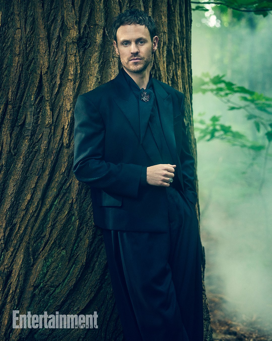 An adult male in a black suit leans against a large tree in a forest with soft, diffused lighting. The man wears a ring and pendant.