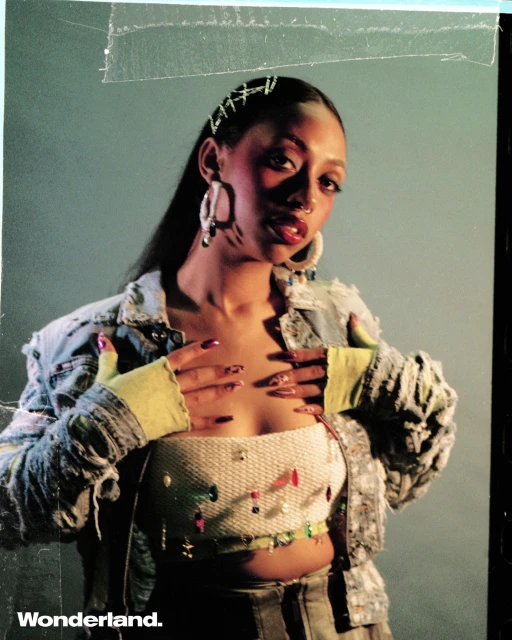 Portrait of a young adult female wearing a denim jacket and a beaded top posing in front of a grey backdrop with dramatic studio lighting.