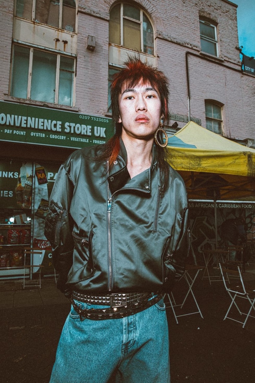 A person with red-tipped hair, wearing a jacket and studded belt, stands in front of a convenience store in an urban setting.