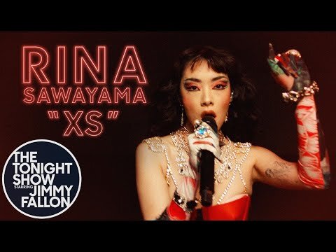 Rina Sawayama sings at The Tonight Show with Jimmy Fallon, wearing a red and white costume, a microphone, and jewelry, performing.
