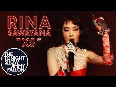 Rina Sawayama sings at The Tonight Show with Jimmy Fallon, wearing a red and white costume, a microphone, and jewelry, performing.