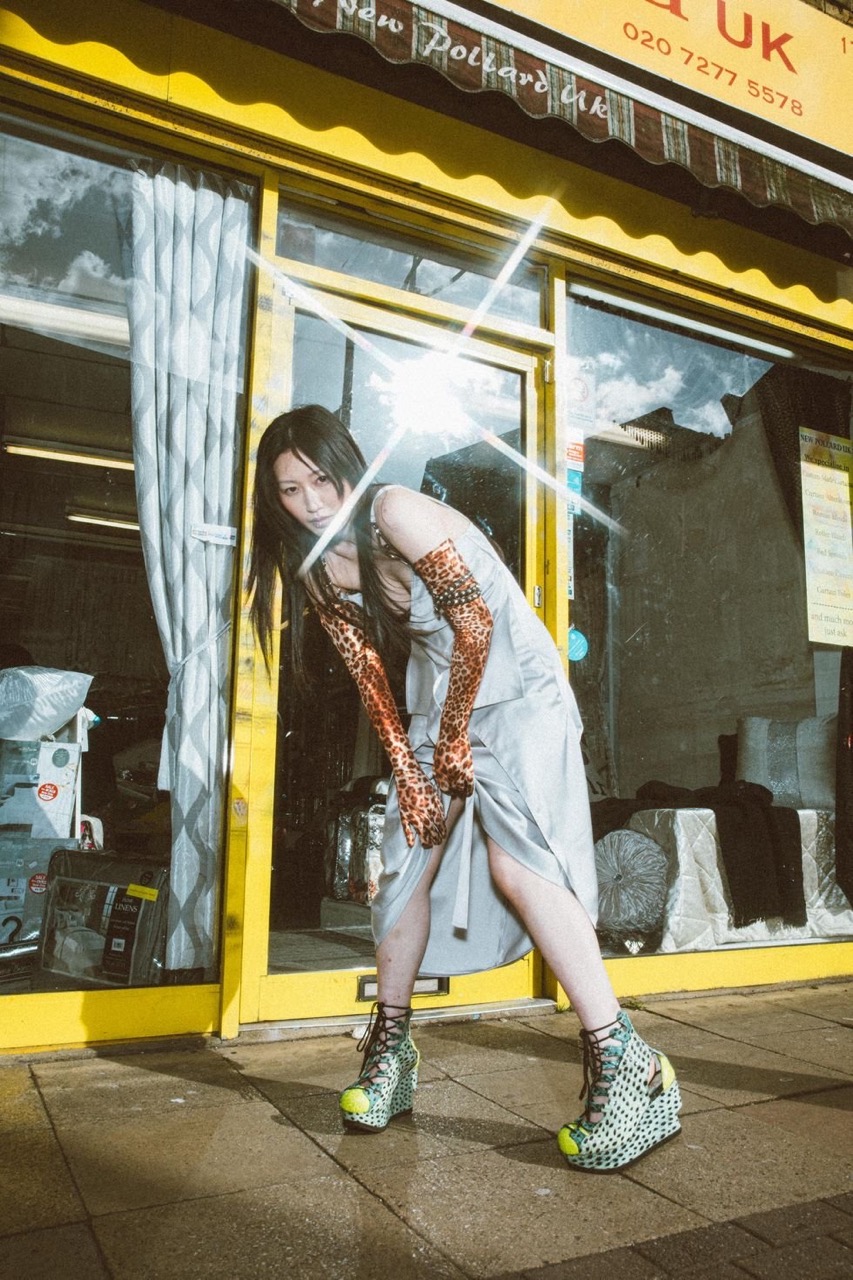 A young woman in a silver dress poses in front of a yellow building wearing leopard-print gloves and platform shoes on a sunny day.
