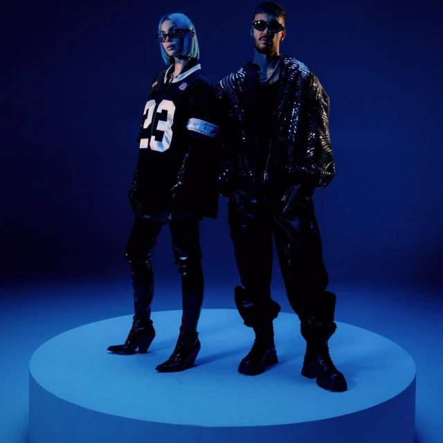 A stylish couple posing on a blue platform in a photo studio, showcasing fashion and style with sports jersey and black sequin jacket.