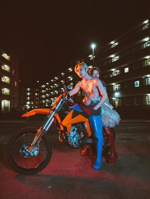 A man and woman sit on an orange motocross bike at night in an urban setting, lit by a street light in a full shot.