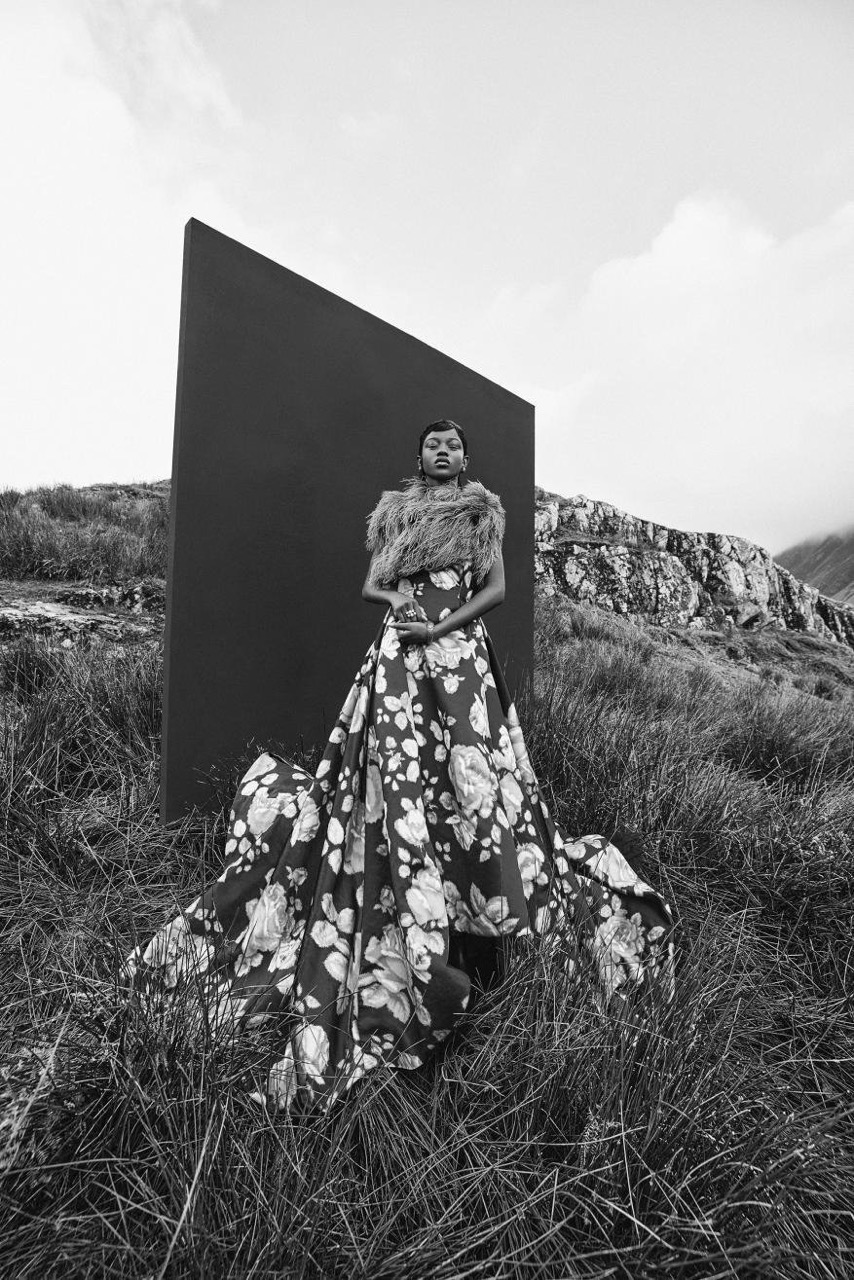Model in a voluminous floral gown and fur stole standing before a large dark rectangular panel in a grassy field