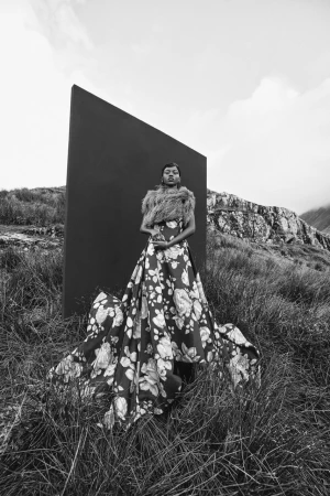 Model in a voluminous floral gown and fur stole standing before a large dark rectangular panel in a grassy field
