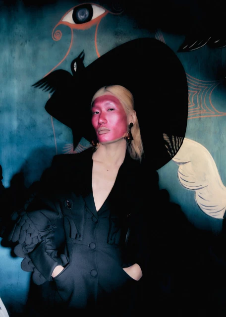Person wearing a black coat and hat posing in front of a painted mural featuring an eye and other abstract elements, indoor shot.