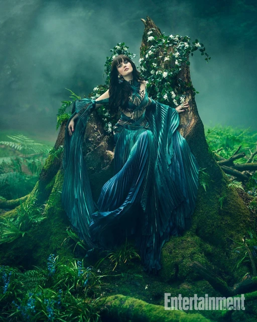 Enchanting woman in teal gown sits on a mossy tree stump in a misty forest with white flowers, creating a magical, fairy-tale aesthetic.