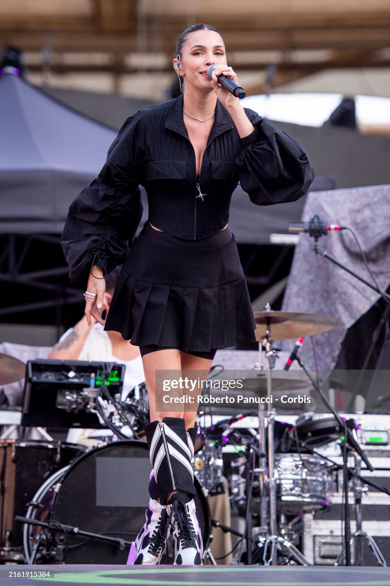 Female singer performs on outdoor stage with microphone, wearing black jacket, pleated mini skirt, and eye-catching boots during concert.