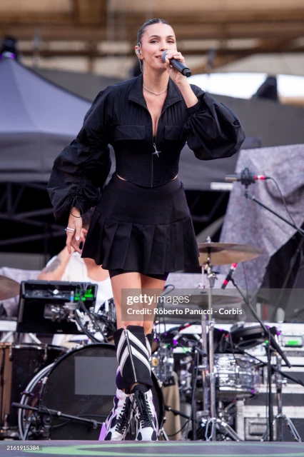 Female singer performs on outdoor stage with microphone, wearing black jacket, pleated mini skirt, and eye-catching boots during concert.
