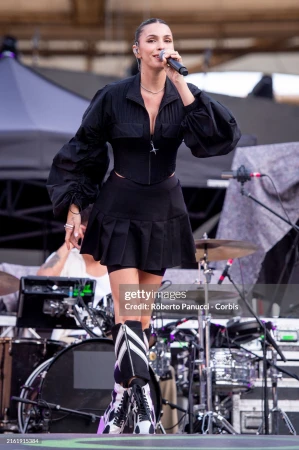 Female singer performs on outdoor stage with microphone, wearing black jacket, pleated mini skirt, and eye-catching boots during concert.