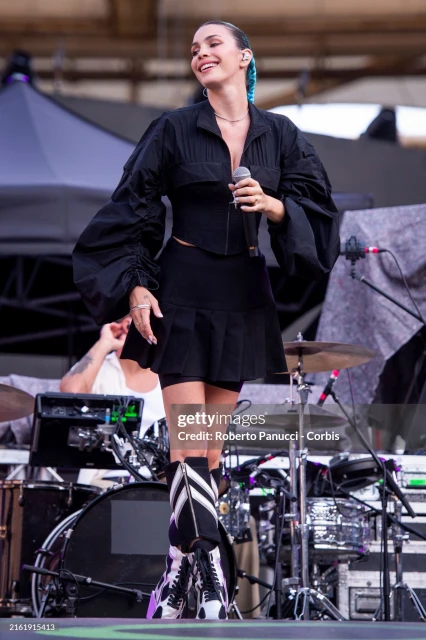 A female singer performs on stage in a black outfit, singing into a microphone, with musical equipment and stage elements in the background.