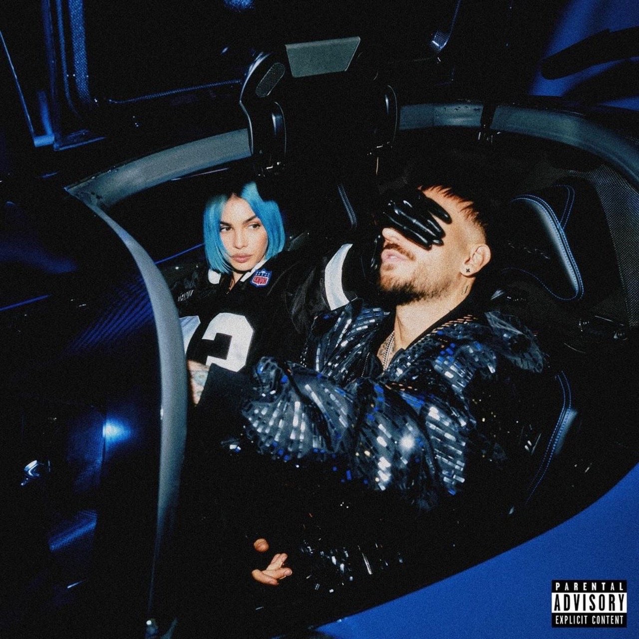Moody indoor shot of a man with gloved hands over eyes and a woman with blue hair inside a car, bathed in blue light.
