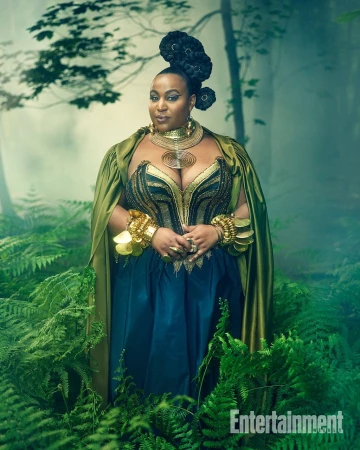Plus size woman in ornate gold jewellery and a green cape standing among lush green ferns in a forest setting.