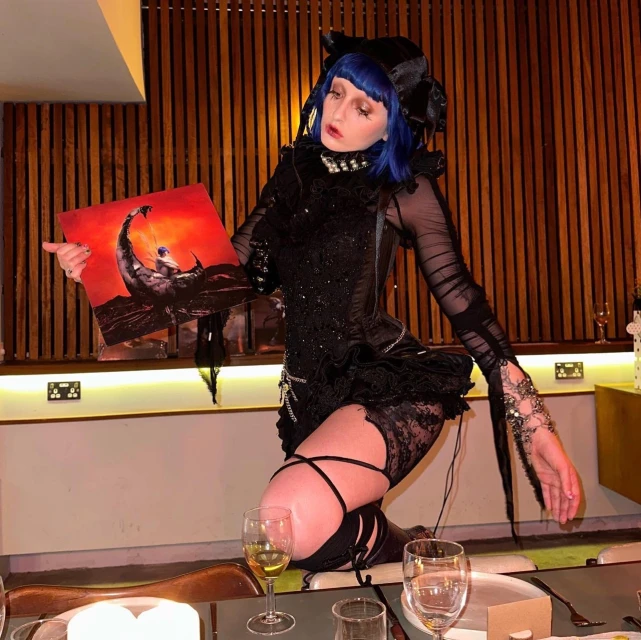 Woman with blue hair and elaborate black lace dress kneels and holds a vinyl record in an indoor setting with dark wood paneling.
