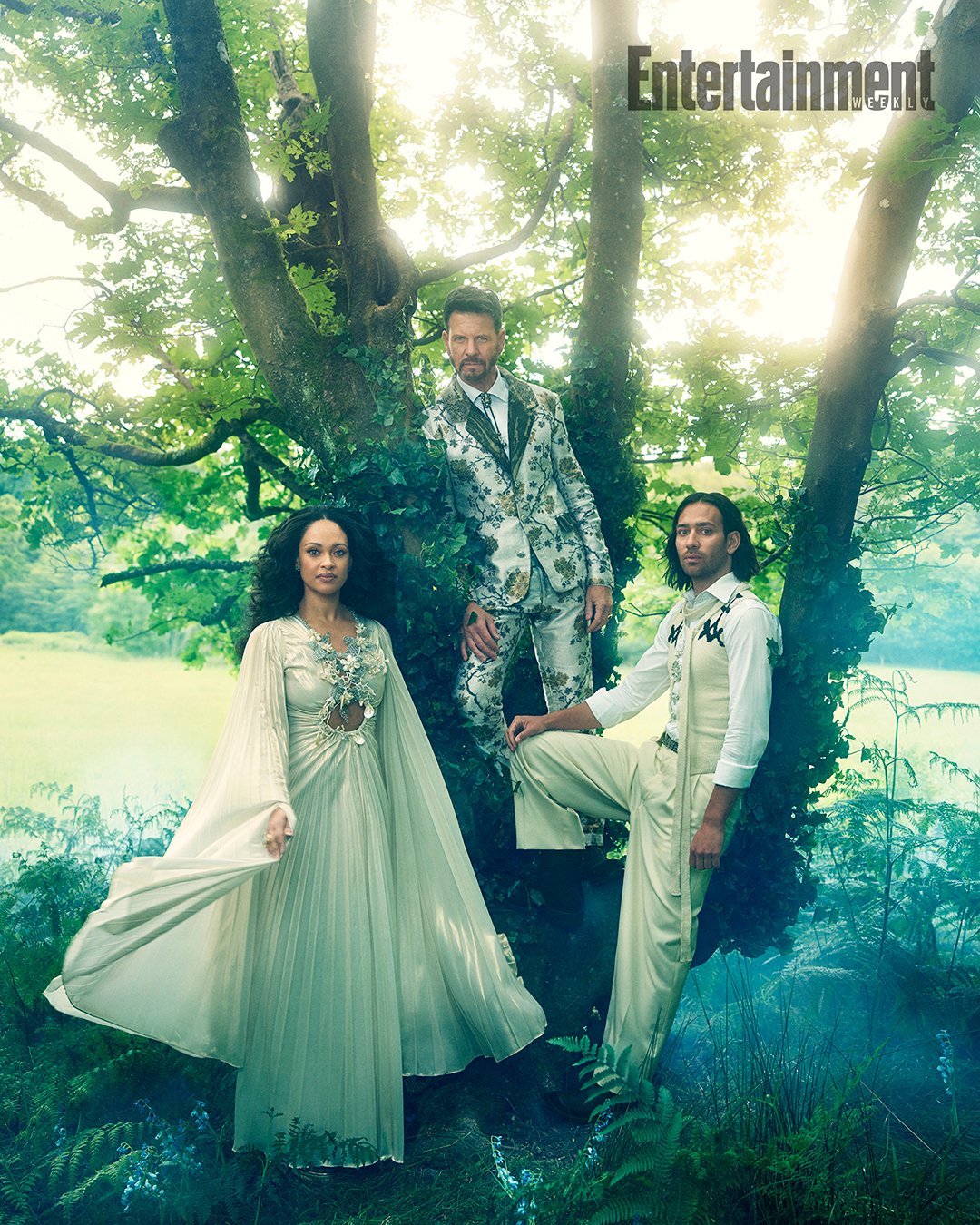 Fashion shot of three models posing in a forest. One woman in a long dress and two men in patterned suits stand among the trees.