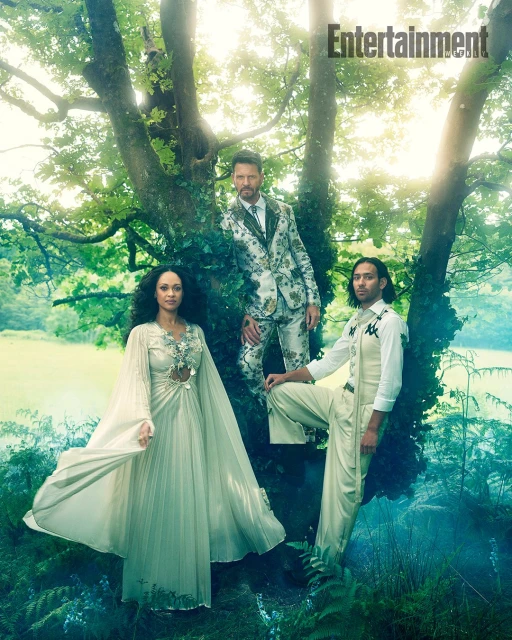 Fashion shot of three models posing in a forest. One woman in a long dress and two men in patterned suits stand among the trees.