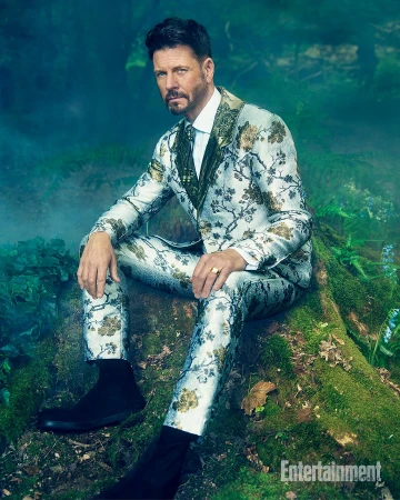 Adult male in a floral suit sitting on a mossy surface in a forest-like studio setting, captured in a medium shot with soft lighting.
