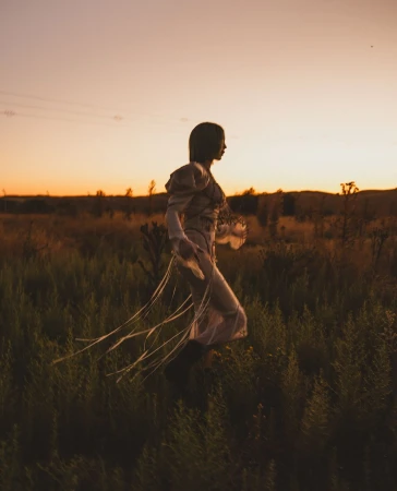 Woman in flowing dress walks through field at sunset. Golden hour lighting, long grass, and gentle breeze create a serene, natural scene.
