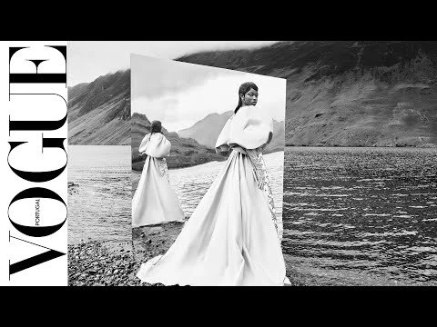 Black and white editorial fashion image featuring a model wearing a sculptural white gown standing before a mirrored panel in a rugged mountainous landscape.