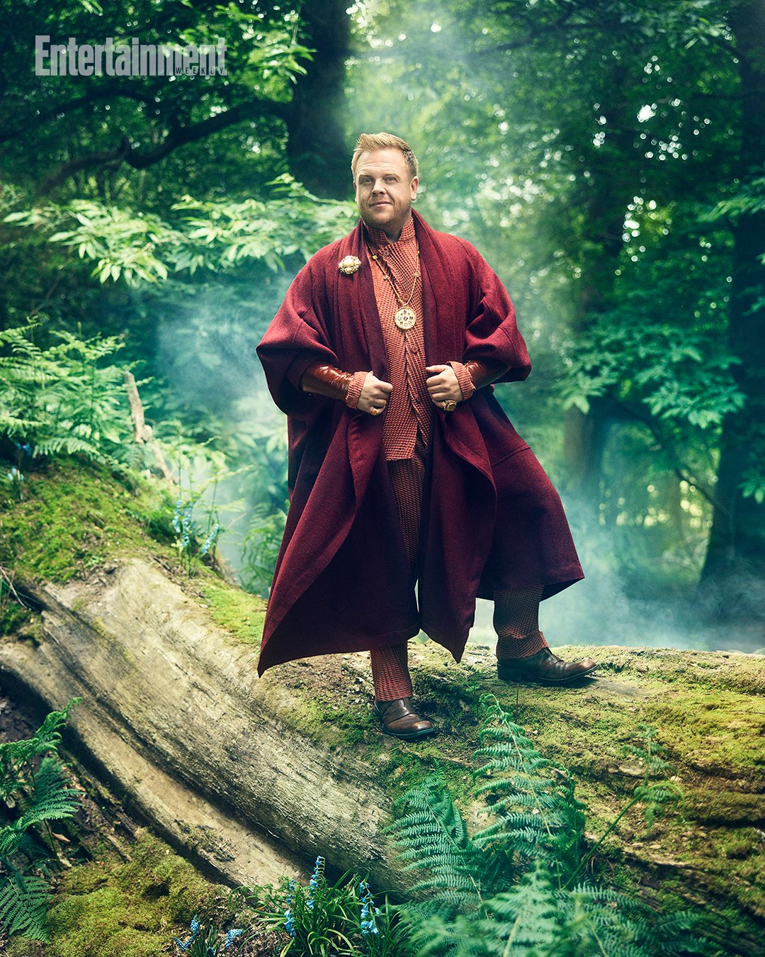 Man in fantasy costume standing on a mossy log in a lush green forest with filtered light and a misty atmosphere, creating a serene scene.