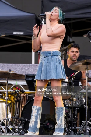 A female singer performs on stage with a microphone, wearing a peach top and denim skirt, backed by a drummer at an outdoor concert.