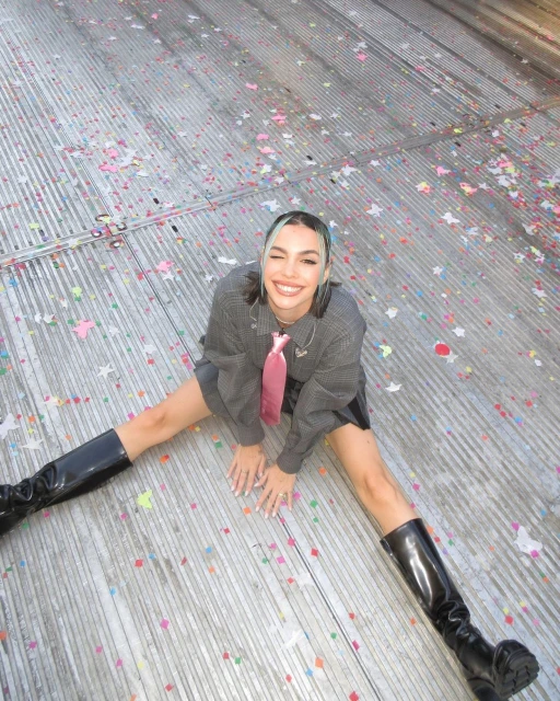 Cheerful woman in black boots and gray outfit strikes a pose on a confetti-covered surface, looking up with a wide smile.