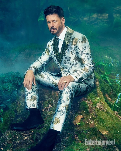 Adult male seated on mossy rock in outdoor forest setting wearing floral-patterned suit and black Chelsea boots in medium shot.