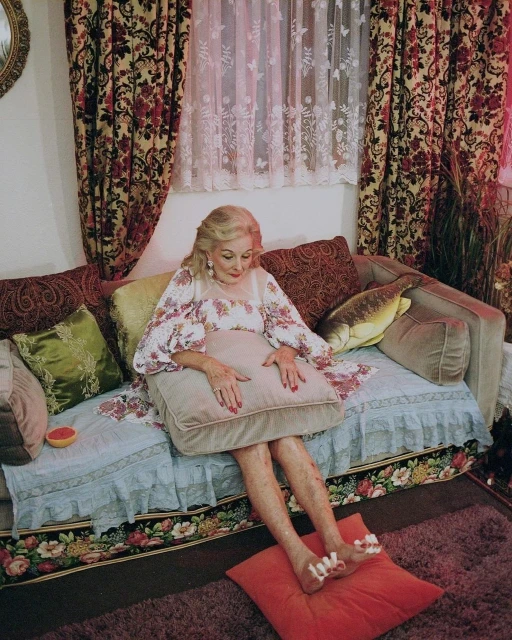 Elderly woman lounges on a floral patterned couch with pillows; she is wearing toe separators for a pedicure in a domestic setting.