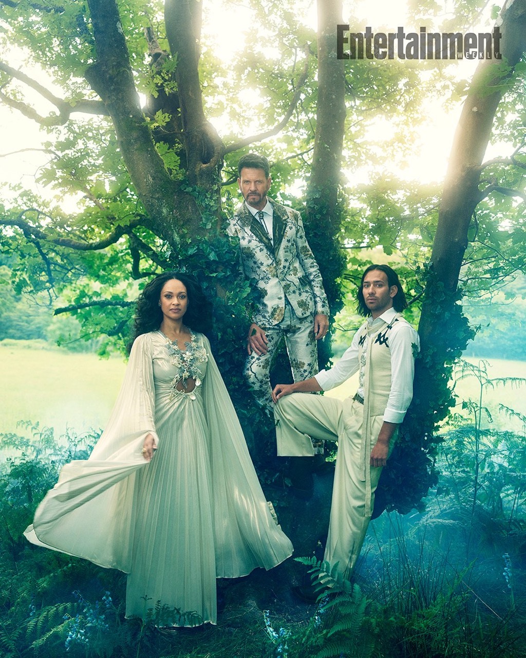 Three actors in floral-patterned costumes pose in a wooded area with greenery and dappled sunlight, promoting a theatrical or entertainment project.