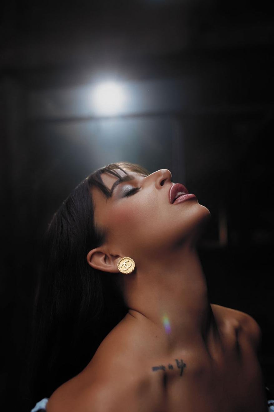Portrait of adult female with bare shoulders and head tilted back, wearing large gold earrings and a dark hairstyle with bangs.