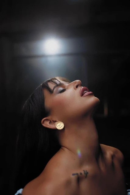 Portrait of adult female with bare shoulders and head tilted back, wearing large gold earrings and a dark hairstyle with bangs. 
