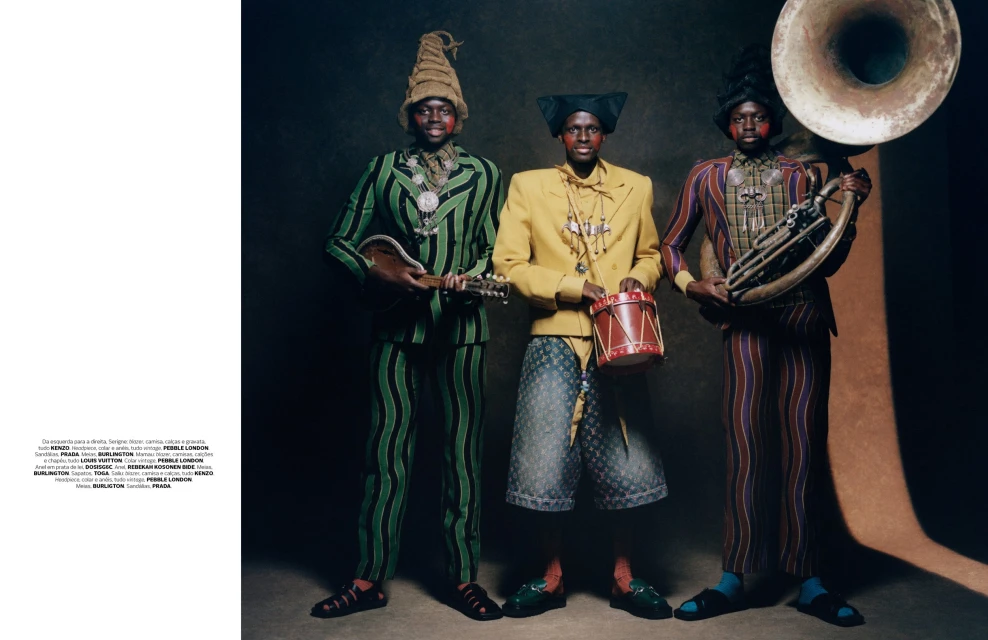 Three men in colorful striped suits pose with musical instruments: a mandolin, a drum, and a tuba against a dark backdrop.