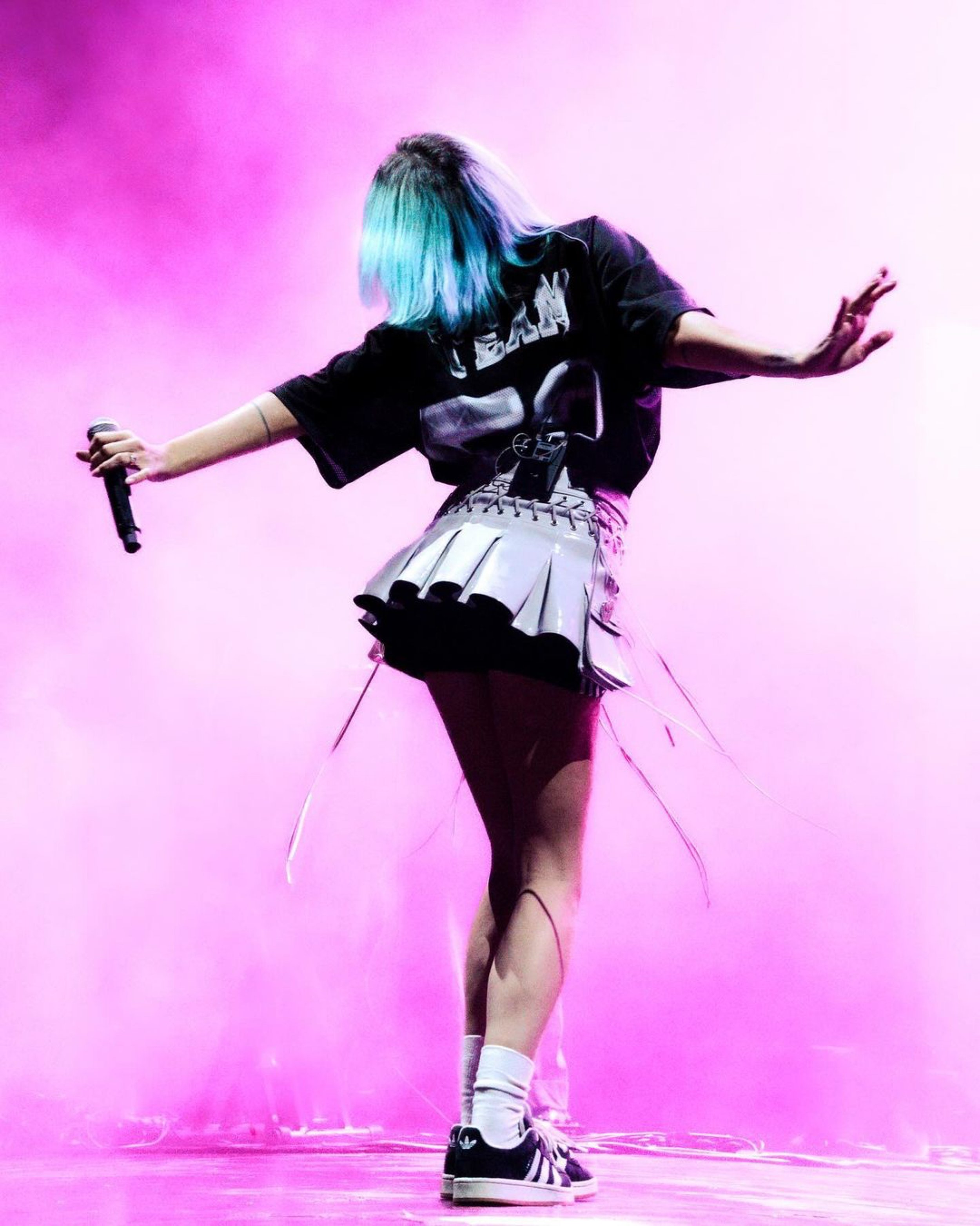 A singer with dyed blue hair in a black t-shirt and white skirt performs on a pink lit stage with a microphone in hand.