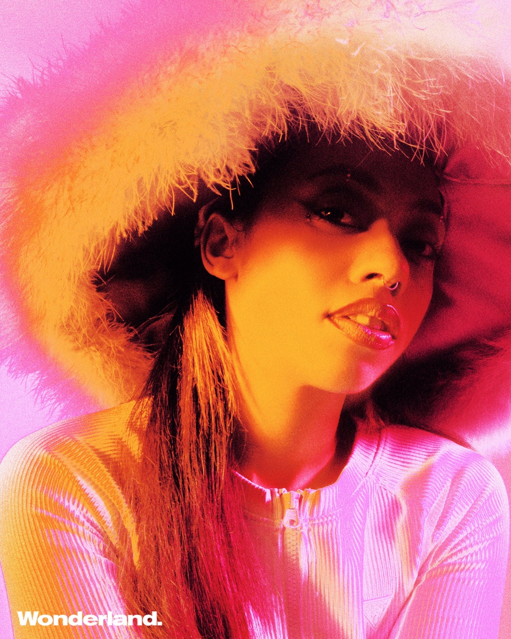 Portrait of a woman wearing a feather hat and pink top with long, red-tinted hair, against a pink backdrop. Wonderland logo visible.