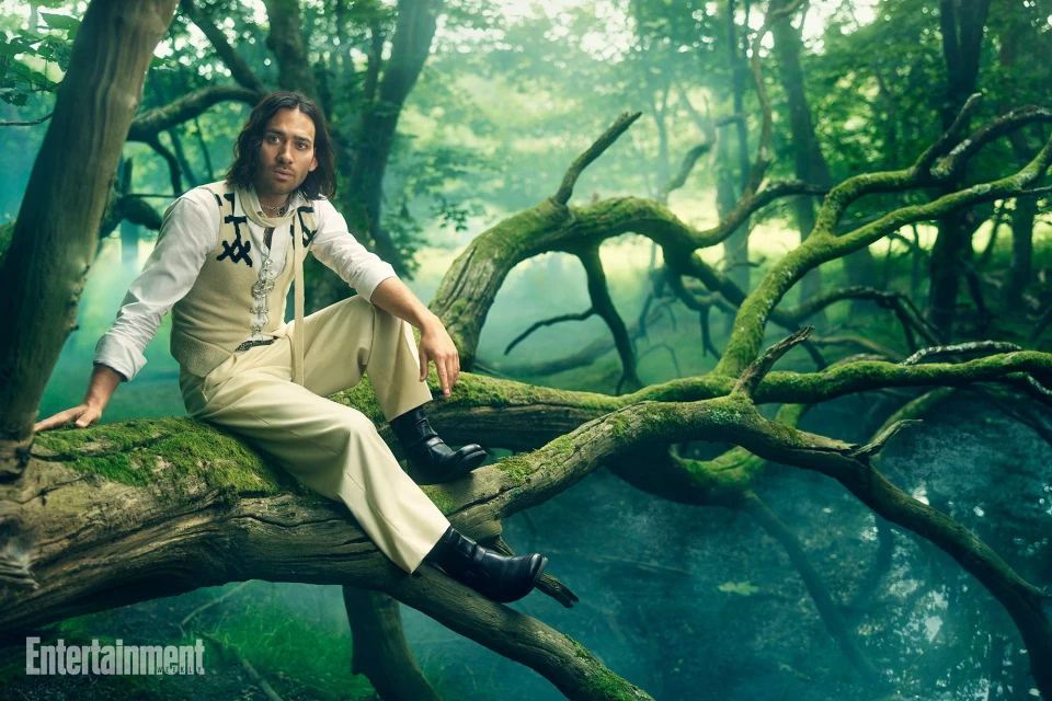 Adult male with long hair sitting on a moss-covered tree branch, wearing a white shirt, vest, black boots, posed in a green forest.