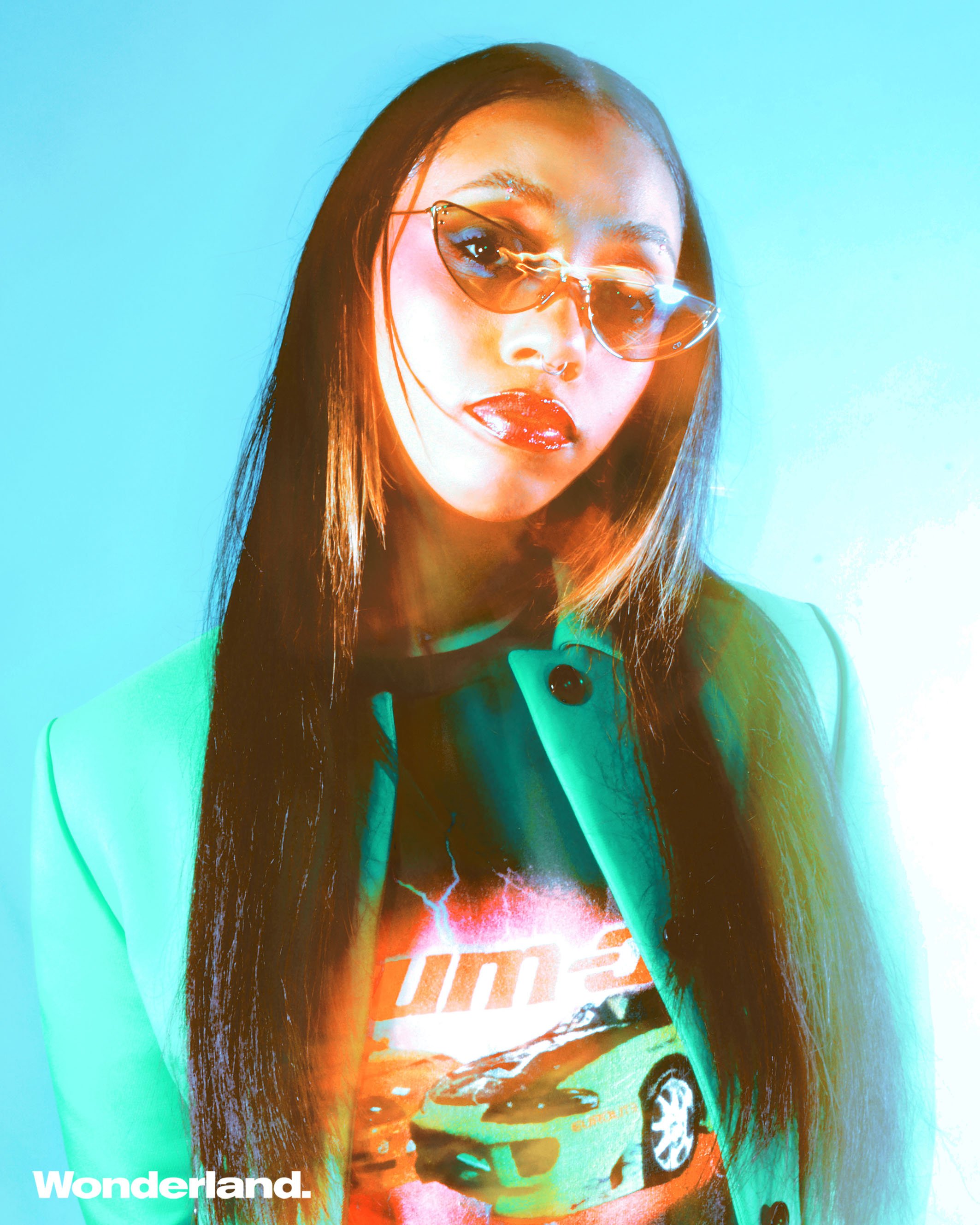Portrait of a young adult woman with long dark hair and cat-eye sunglasses wearing a jacket and a graphic t-shirt against blue background.