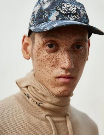 Adult male portrait wearing a beige turtleneck sweater and a blue tiger patterned baseball cap with a designer logo on the front.