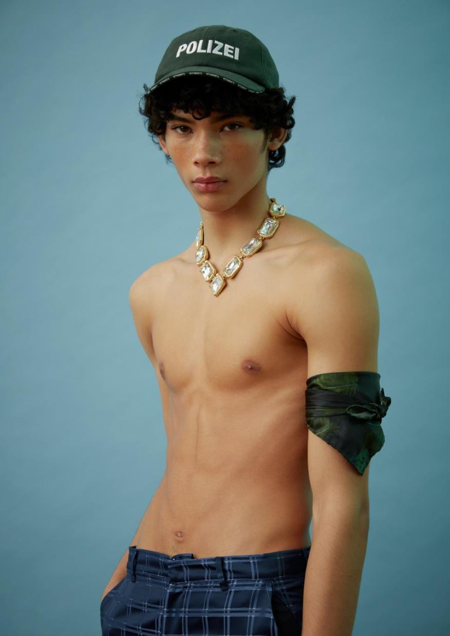 A young adult with fair skin poses shirtless against a blue background, wearing a cap, statement necklace, arm band, and plaid pants.