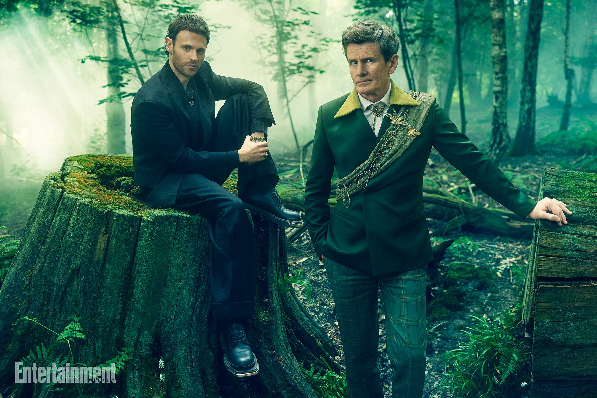 Two men pose in a forest setting; one sits on a mossy tree stump, wearing black, while the other stands in green attire.