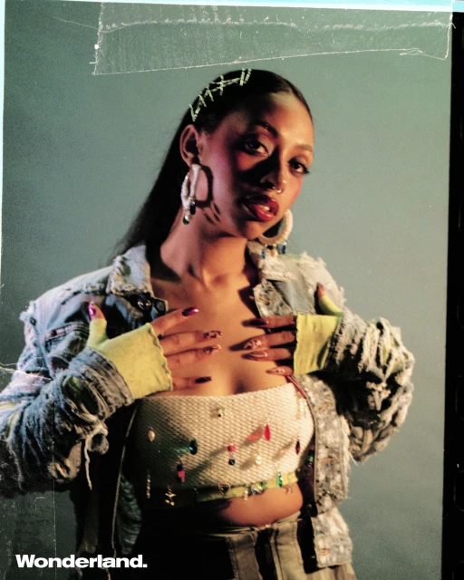 Portrait of a young woman with long dark hair, wearing a beaded crop top and denim jacket, posing in front of a muted green background.